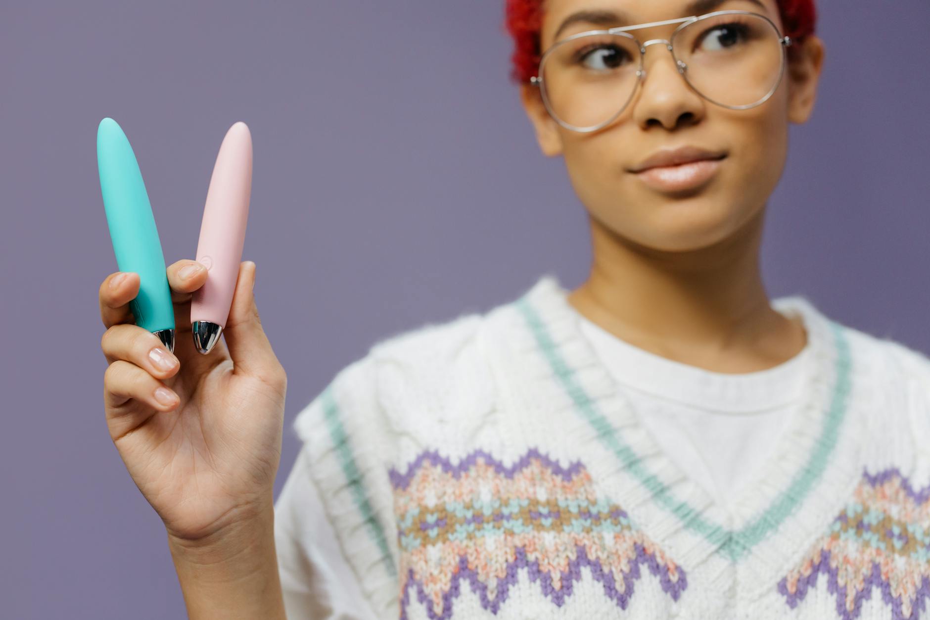 Woman with eyeglasses holding colorful vibrators against a purple background.