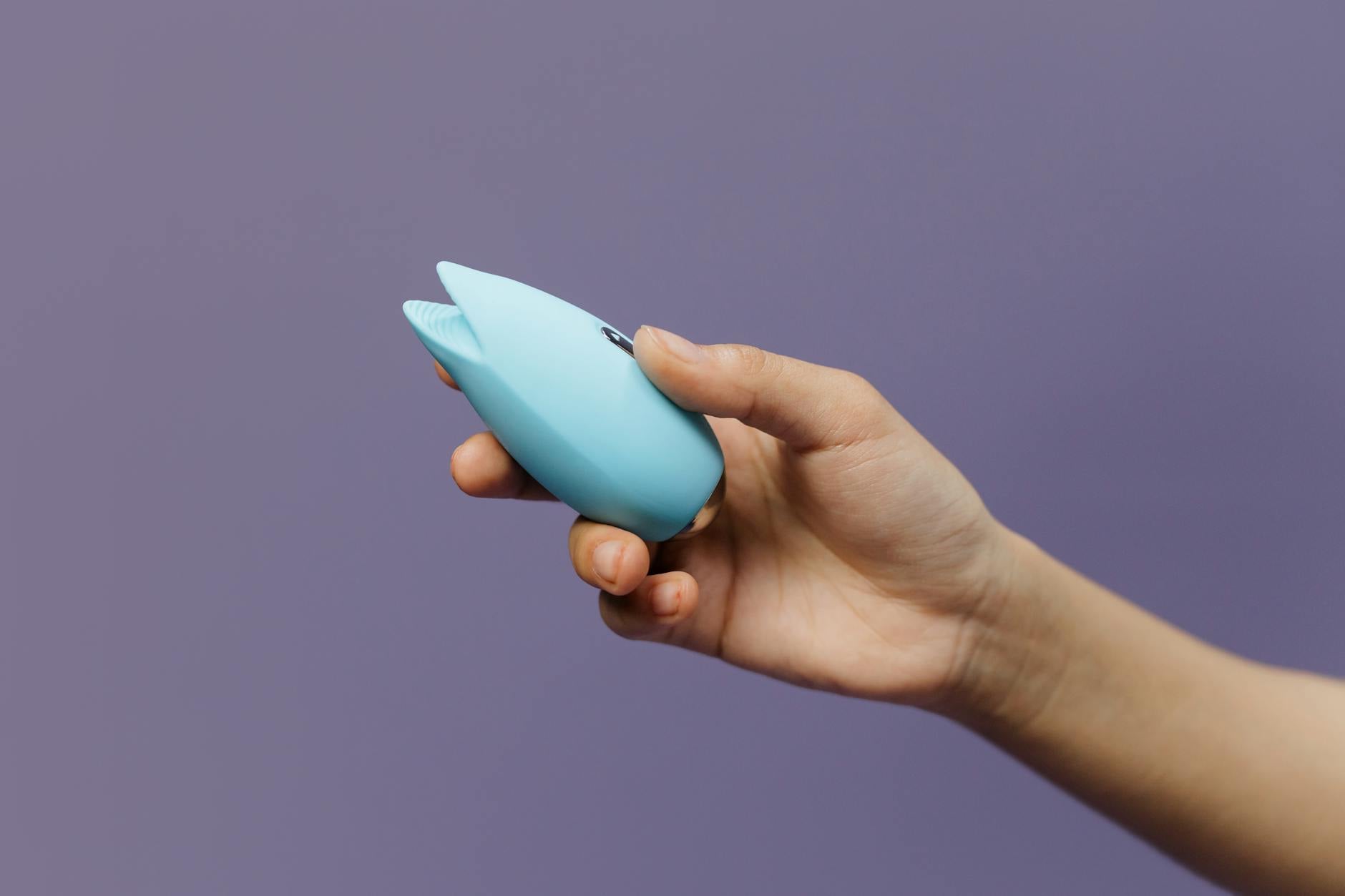 A blue silicone clitoral vibrator held in hand against a purple background, representing accessible pleasure and self-care.